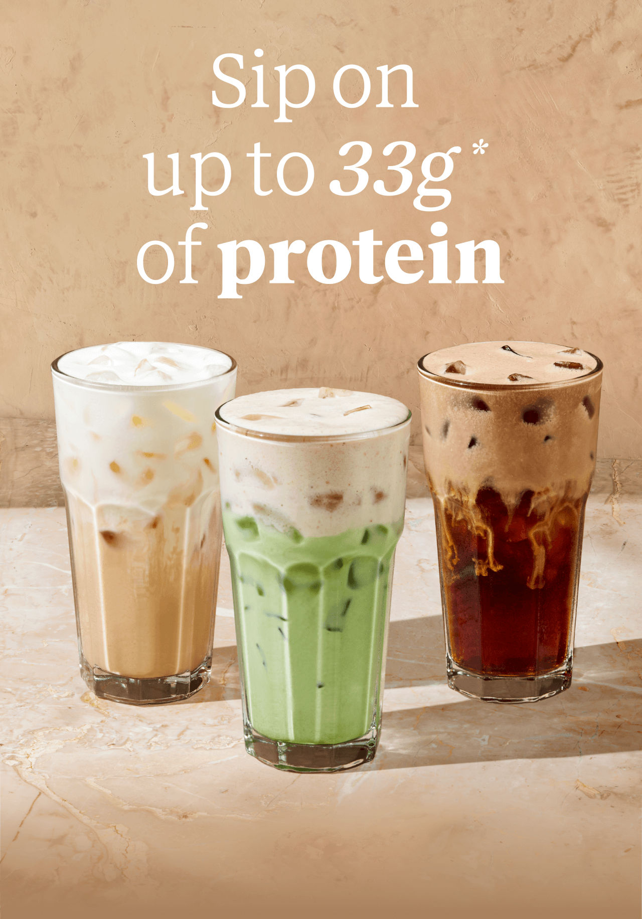 Sip on up to 33g* of protein | We see a refreshing iced vanilla latte on the left beige in colour, next to a milky-green iced banana matcha in the centre, and a rich brown chocolate cream cold brew on the right. All three drinks are topped with a creamy protein foam against a warm beige background.
