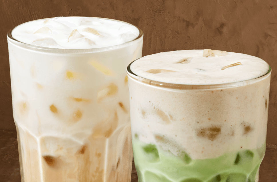 We see two iced drinks – the one on the left is a refreshing vanilla beverage beige in colour, next to a milky-green iced banana matcha. Both are topped with a creamy protein foam against a brown background.