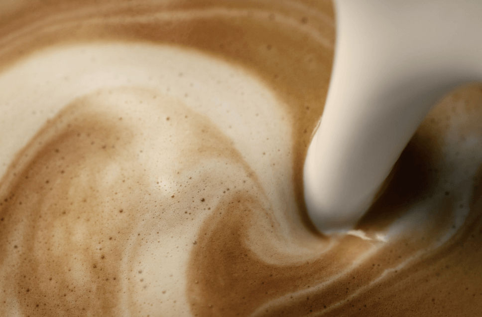 A close-up, bird's eye view of creamy protein milk being poured into a creamy latte. There is a blend of golden espresso tones with milk cascading together.