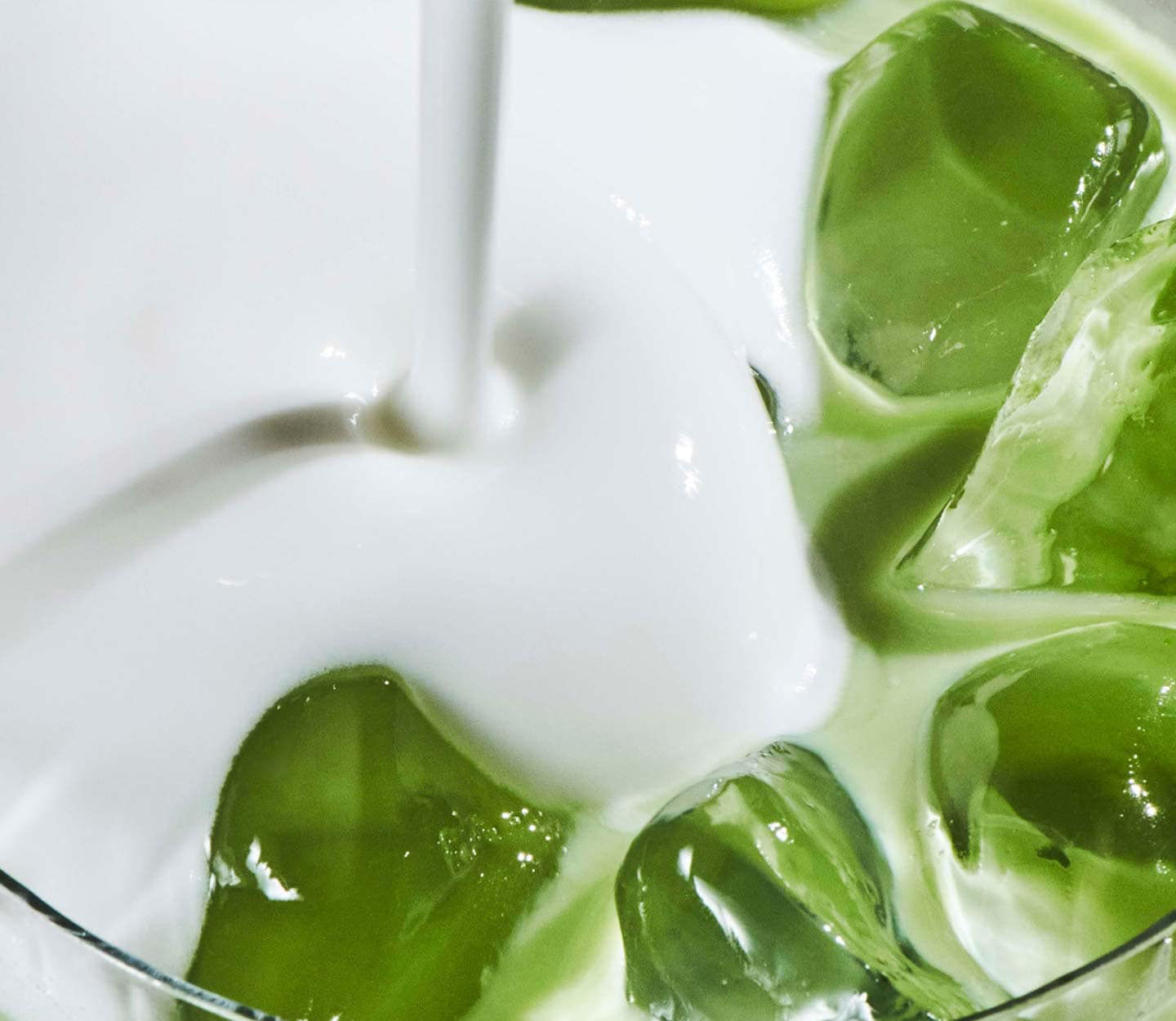 Close-up of milk being poured over a green iced beverage, swirling around large ice cubes in a glass.