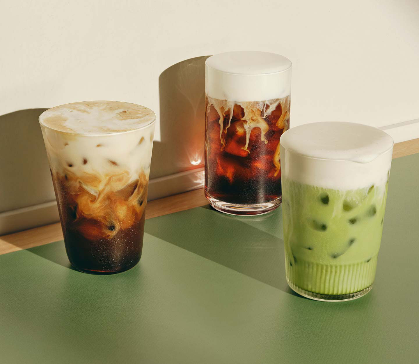 Three handcrafted iced beverages on a tabletop, including a green iced drink and two dark coffee drinks topped with creamy protein foam.