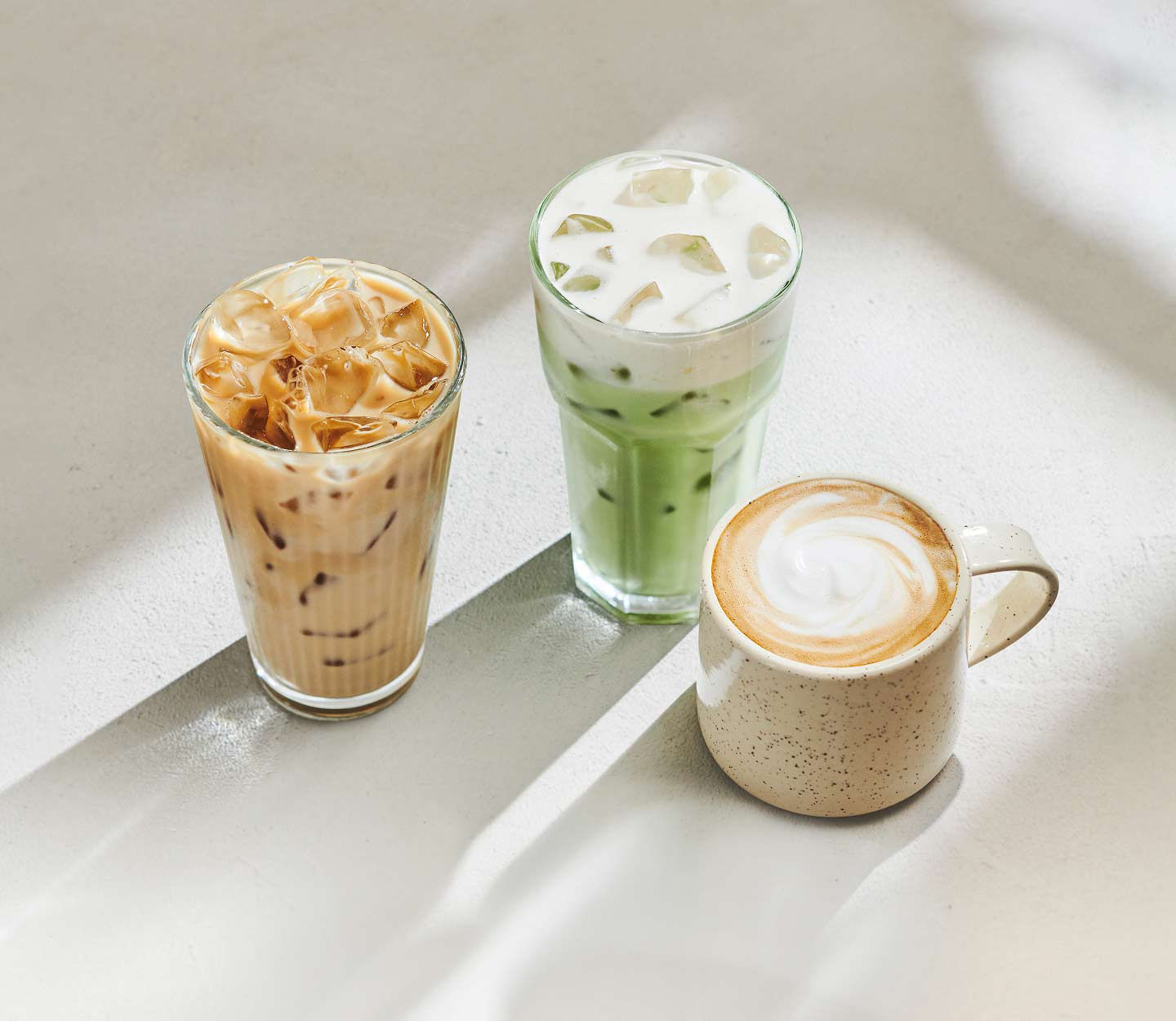 Three protein beverages arranged on a light tabletop, including an iced latte, a green iced drink and a hot latte with foam art.