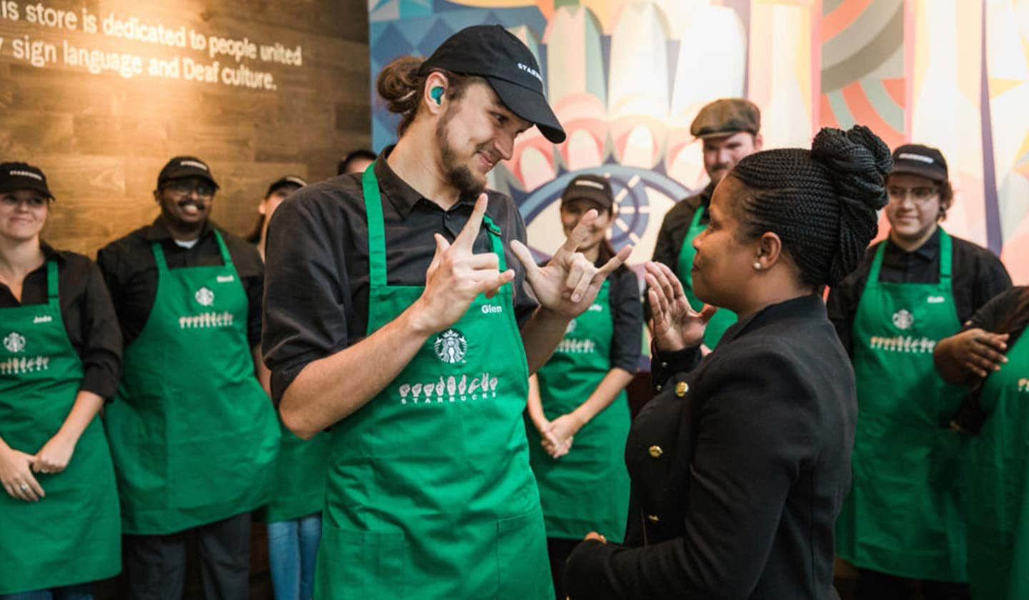 Photo d'un homme et d'une femme qui communiquent en langue des signes. Ils portent un tablier vert Starbucks sur lequel est indiqué qu'ils connaissent la langue des signes et sont entourés d'autres employés.
