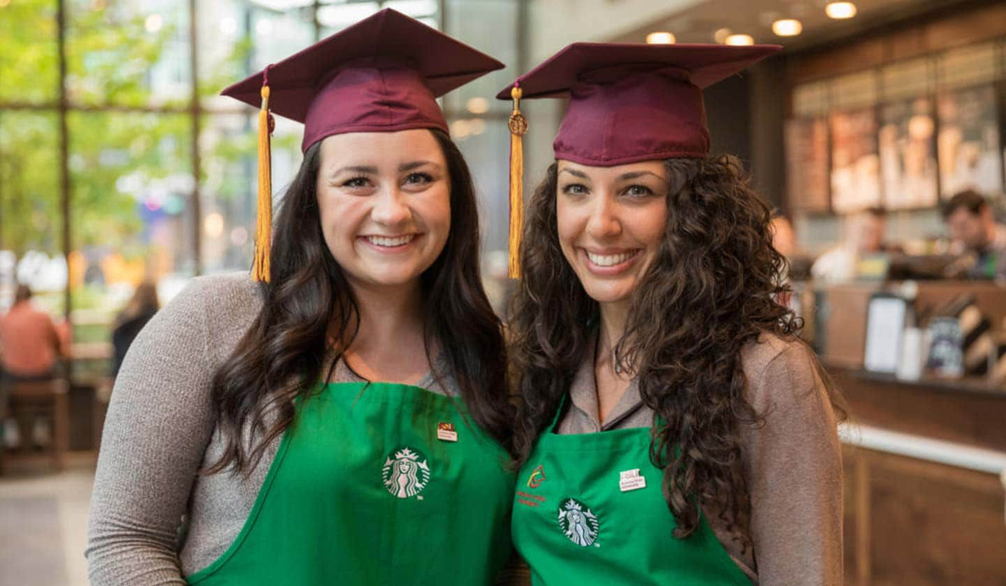 Deux femmes dans un magasin Starbucks sourient pour la caméra. Elles portent le tablier vert ainsi qu'un chapeau de remise des diplômes rouge bordeaux.