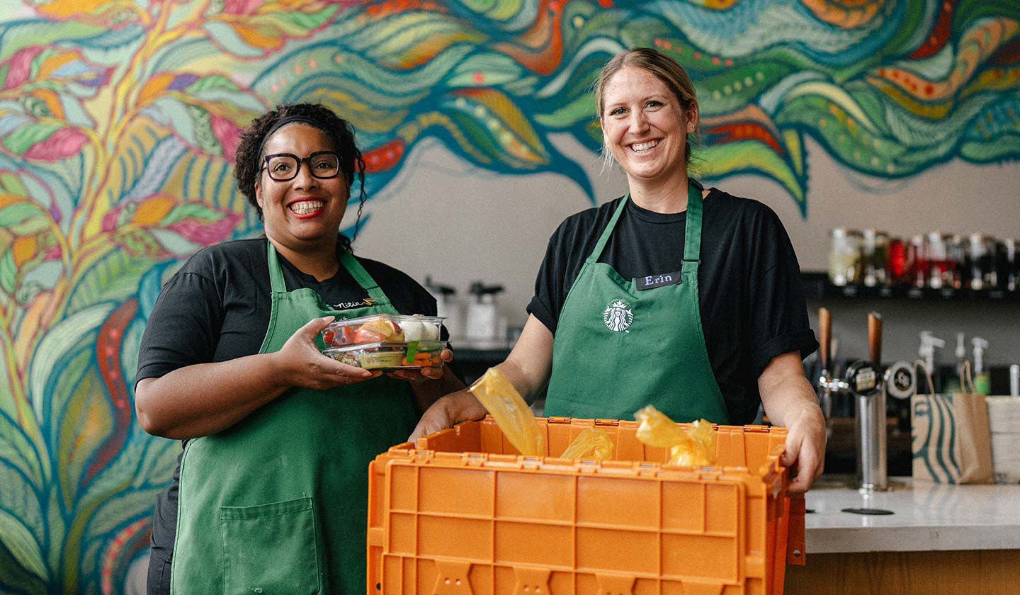 Deux employés Starbucks dans un magasin Starbucks sourient pour la caméra tout en montrant une caisse jaune d'aliments emballés.